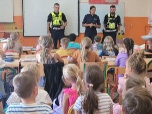 W klasie szkolnej uczniowie siedzą na pogadance. Na środku stoi 2 policjantów i 1 policjantka, którzy prowadzą zajęcia.