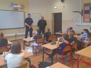 W klasie szkolnej uczniowie siedzą na pogadance. Na środku stoi 2 policjantów, którzy prowadzą zajęcia.