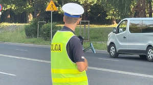 Na zdjęciu policjant ruchu drogowego wraz z urządzeniem do pomiaru prędkości. W tle jedzie samochód dostawczy
