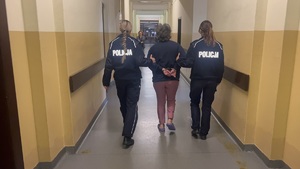 Na korytarzu w Komendzie Miejskiej Policji w Gliwicach widać jak dwie funkcjonariuszki prowadzą w kajdankach zatrzymaną.