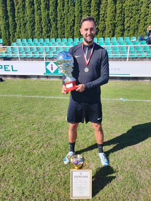 Policjant ubrany w strój sportowy stojący z pucharem na boisku trawiastym.