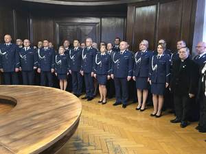 Wzdłuż ściany stoją umundurowane policjantki i policjanci w umundurowaniu galowym podczas uroczystości.