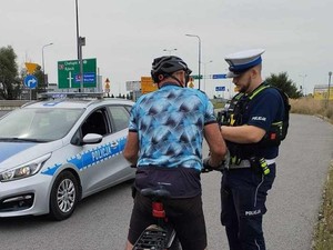 Na zdjęciu rower i zatrzymany rowerzysta podczas kontroli prowadzonej przez policjanta ruchu drogowego. W tle stoi radiowóz.
