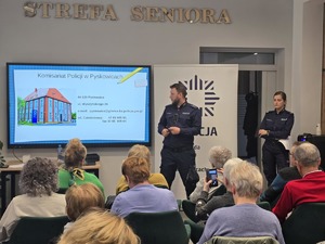 Na zdjęciu policjant prowadzący debatę społeczną stojący przy tablicy edukacyjnej. Na sali siedzą mieszkańcy miasta.