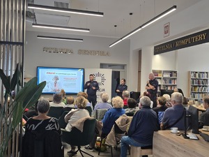 Na zdjęciu policjant prowadzący debatę społeczną stojący przy tablicy edukacyjnej. Na sali siedzą mieszkańcy miasta.