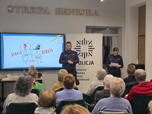 Na zdjęciu policjant prowadzący debatę społeczną stojący przy tablicy edukacyjne - obok niego policjantka. Na sali siedzą mieszkańcy miasta.