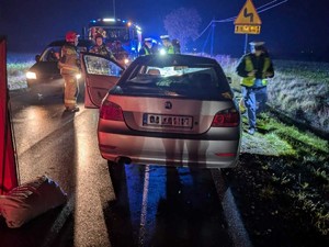 Na jezdni po zmierzchu stoi pojazd po potrąceniu ze skutkiem śmiertelnym pieszego. W tle widać wóz straży Pożarnej oraz strażaków
