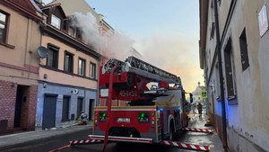 Pojazd straży pożarnej stojący na jezdni podczas akcji gaszenia pożaru. Z okna budynku widać wydobywające się kłęby dymu.