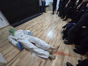 Zajęcia dla uczniów klasy policyjnej prowadzi technik kryminalistyki. Dookoła niego w pokoju stoją uczniowie. Na podłoże leży manekin symulujący ofiarę.