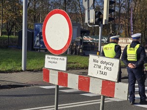 Na zdjęciu policjanci ruchu drogowego podczas zabezpieczenia ruchu w rejonie cmentarza. Stoją przy znakach drogowych na jezdni.