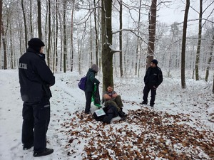 W lesie po drzewem siedzi rozebrany człowiek. Pomocy udzielają jej harcerze jej pomocy. Obok stoją policjanci.