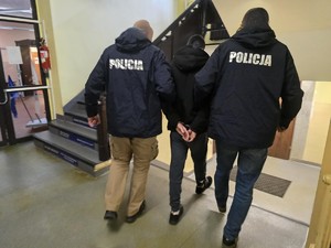 policjanci w granatowych kurtkach z napisem na plecach POLICJA prowadzą zatrzymanego