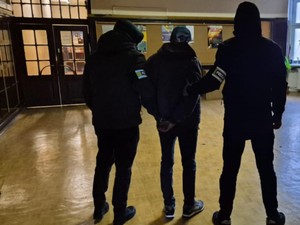 W holu dwóch mężczyzna, którego pilnuje dwóch nieumundurowanych policjantów.