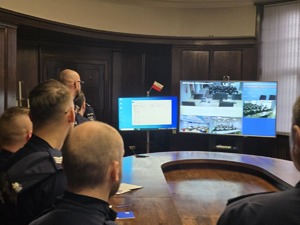 Na końcu sali stoją monitory. Na środku okrągły stół, przy którym stoją słuchacze.