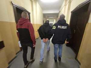 Na zdjęciu widoczni dwaj nieumundurowani funkcjonariusze policji prowadzący zatrzymanego mężczyznę w budynku policji. Zatrzymany ma założone kajdanki na ręce.