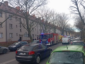 Na zdjęciu na ulicy stoi straż pożarna oraz karetka pogotowia i radiowóz Policji. W tle budynki mieszkalne. Ulicą przemieszczają się pojazdy