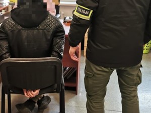 Na zdjęciu siedzi podejrzany, obok stoi policjant w cywilnym ubraniu