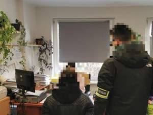 Zatrzymany siedzi przy biurku, obok stoi nieumundurowany policjant.