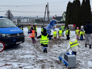 Dzieci w kamizelkach odblaskowych biegają na dworze. Na zdjęciu stoi radiowóz policyjny, a nieopodal Sznupek.