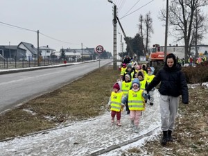 Grupa dzieci w kamizelkach odblaskowych idą chodnikiem z opiekunką