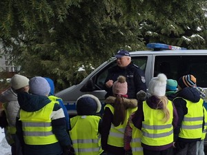 Grupa dzieci w kamizelkach odblaskowych stoi koło radiowozu Policji. Policjant przeprowadza pogadankę dla dzieci.
