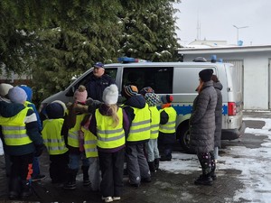 Grupa dzieci w kamizelkach odblaskowych stoi koło radiowozu Policji. Policjant przeprowadza pogadankę dla dzieci.