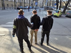 Widok od tyłu zatrzymanego i dwóch nieumundurowanych policjantów stojących przed przejściem dla pieszych