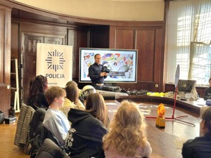 Przy stole siedzą uczniowie. Zajęcia prowadzi policjant ruchu drogowego