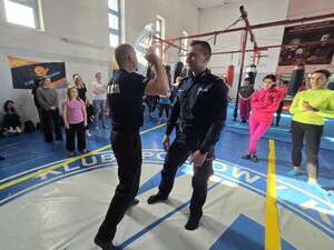 Dwóch policjantów stojących na macie pokazują samoobronę. Dookoła nich stoją kobiety