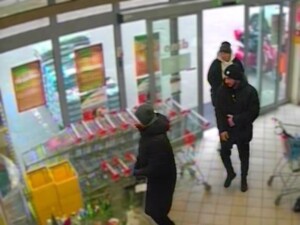 Na zdjęciu drzwi wejściowe do sklepu. Wchodzi dwóch mężczyzn i jedna kobieta. W tle stoją wózki sklepowe.