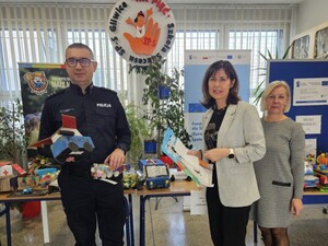 Policjant w mundurze i dwie kobiety stoją przed stołami na których leżą prace plastyczne