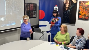 na zdjęciu widzimy jak jedna z seniorek przekazuje prowadzącej bukiet wiosennych kwiatów.