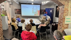 na zdjęciu widzimy seniorów siedzących przy stole i słuchających prelekcji przedstawiciela banku.