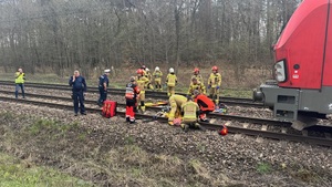 Na torowisku przy czerwonej lokomotywie widzimy wszystkie służby pracujące przy poszkodowanej leżącej na noszach na torach.