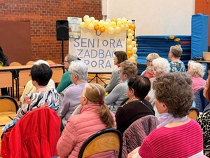 Na sali gimnastycznej na krzesłach siedzą seniorzy. W tle tablica z napisem "O seniora-zadbać pora"