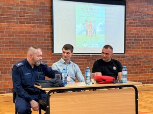 Przy stole siedzi policjant, strażak i lekarz. W tle na ścianie widać ekran projekcyjny