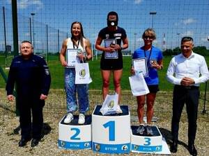 Na podium zwycięski zawodniczki biegu. Po obu stronach stoją mężczyźni, w tym jeden z nich jest w mundurze
