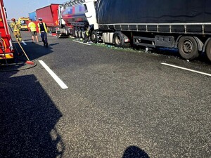 Na autostradzie stoją pojazdy ciężarowe oraz karetka pogotowia. Jezdnią idzie policjant . Miejsce wypadku drogowego.