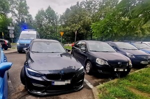 Na parkingu stoją samochody. W tle radiowóz oznakowany Polilicji
