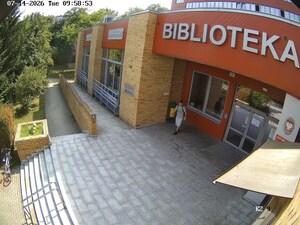 Mężczyzna w czapce bejsbolówce i koszulce i w spodenkach stojący przed budynkiem. Na plecach ma plecak.