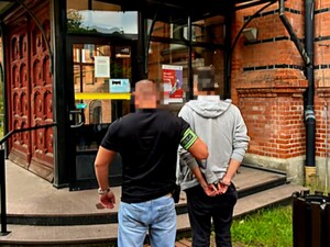 Nieumundurowany policjant prowadzi zatrzymanego w kierunku drzwi wejściowych do budynku.