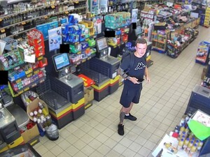 Młody mężczyzna stoi między półkami w sklepie. Ubrany w koszulkę granatową i krótkie spodenki.